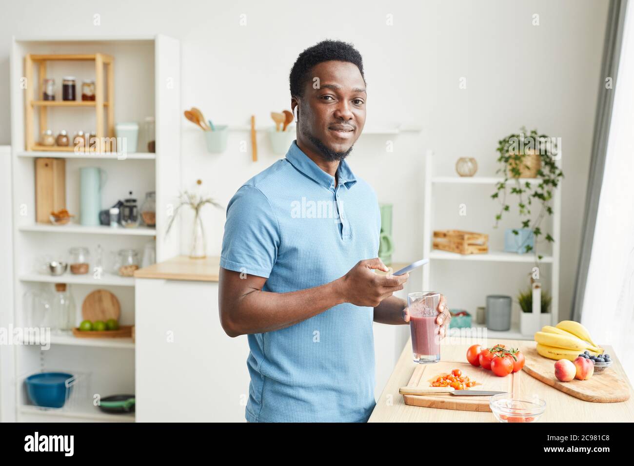 Portrait of African young man smiling at camera while drinking juice and using his mobile phone in the domestic kitchen Stock Photo