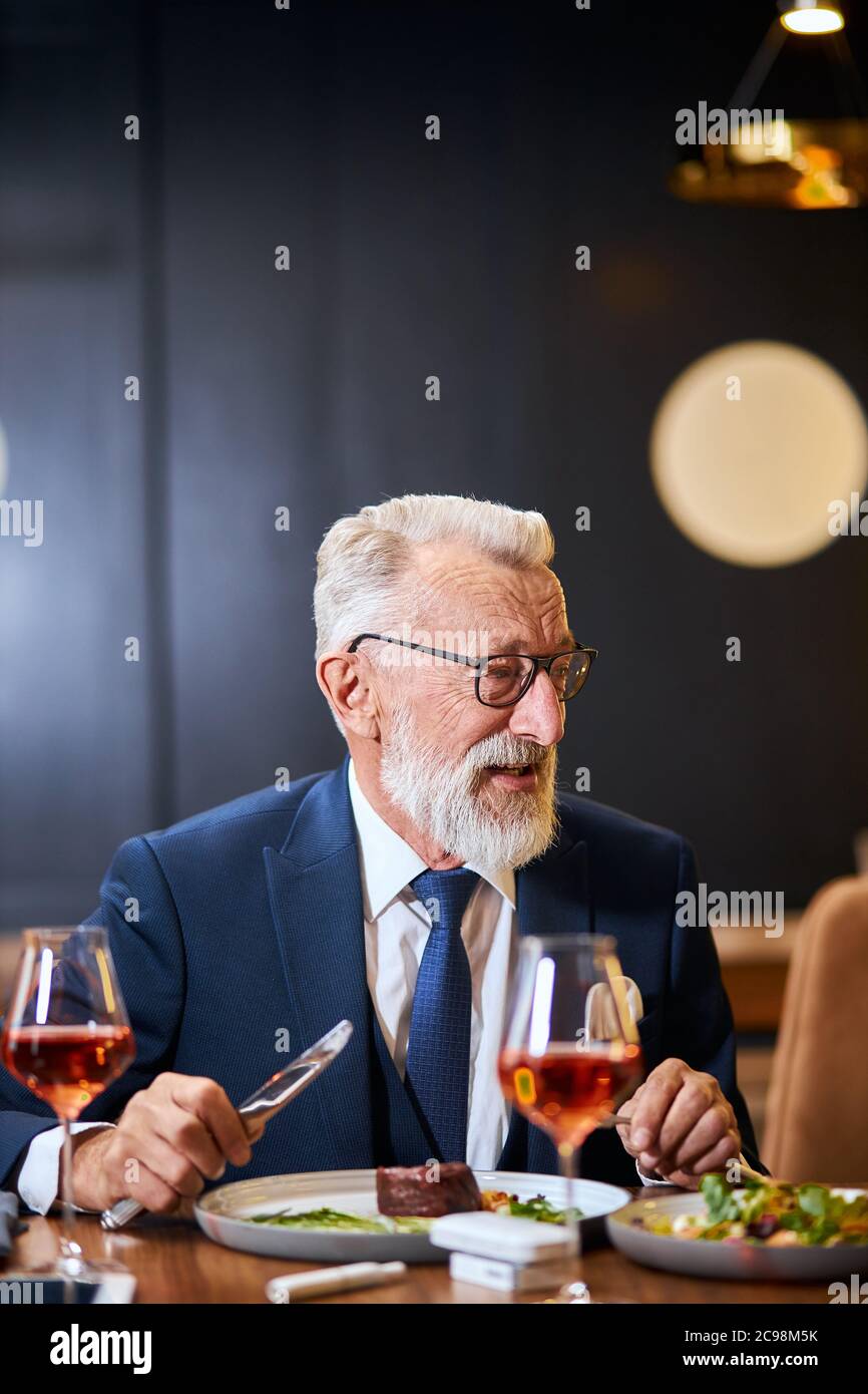 Bearded elderly businessman in glasses on a meeting in a restaurant, having a conversation with business partner and taking a meal. Man eats from beau Stock Photo