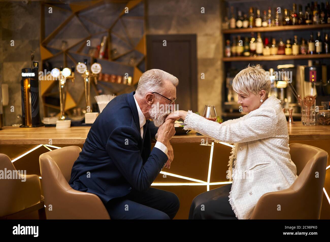 Handsome man in beautiful tuxedo and woman in white blazer sit in expensive beautiful restaurant. Male kissing hand and declare his love. Romantic ima Stock Photo