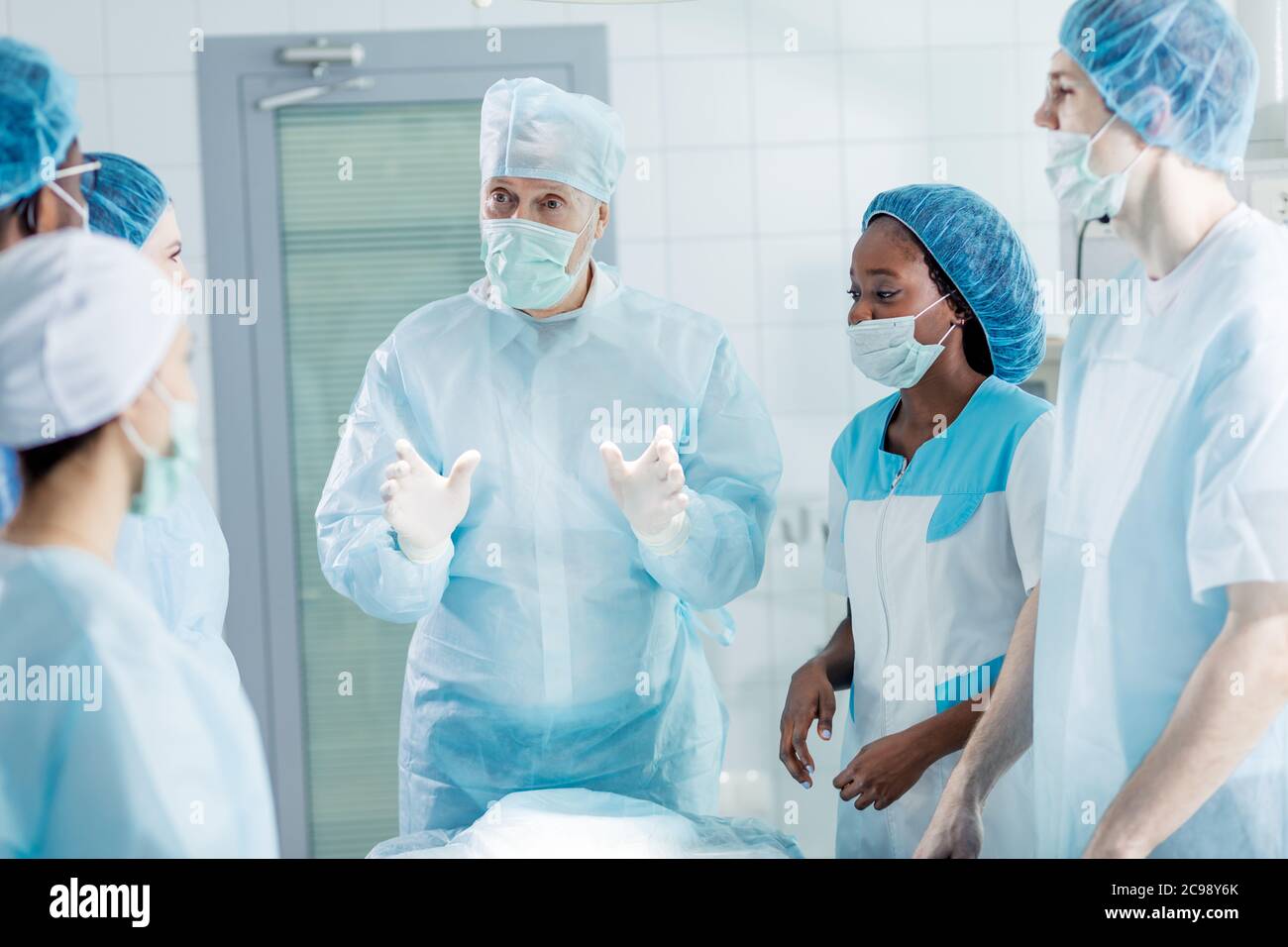 clever surgeon in mask and cap explaining something to his co-workers who are listerning to him very attentively. Stock Photo