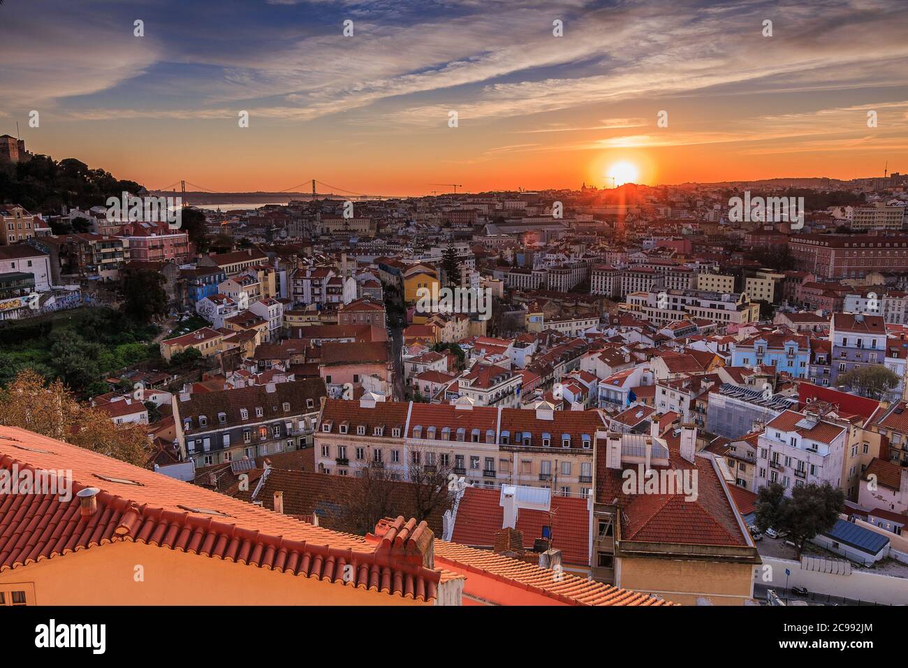 Sunset over the rooftops of the city of Lisbon. Skyline of the old town of the Portuguese capital. Clouds and sun rays in the evening. Stock Photo