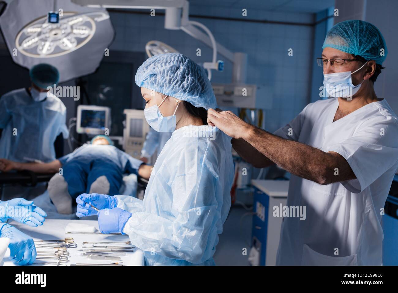 surgery, medicine and people concept - Male doctor assisting female surgeon and helping her with gloves and protective wear in operating room at hospi Stock Photo