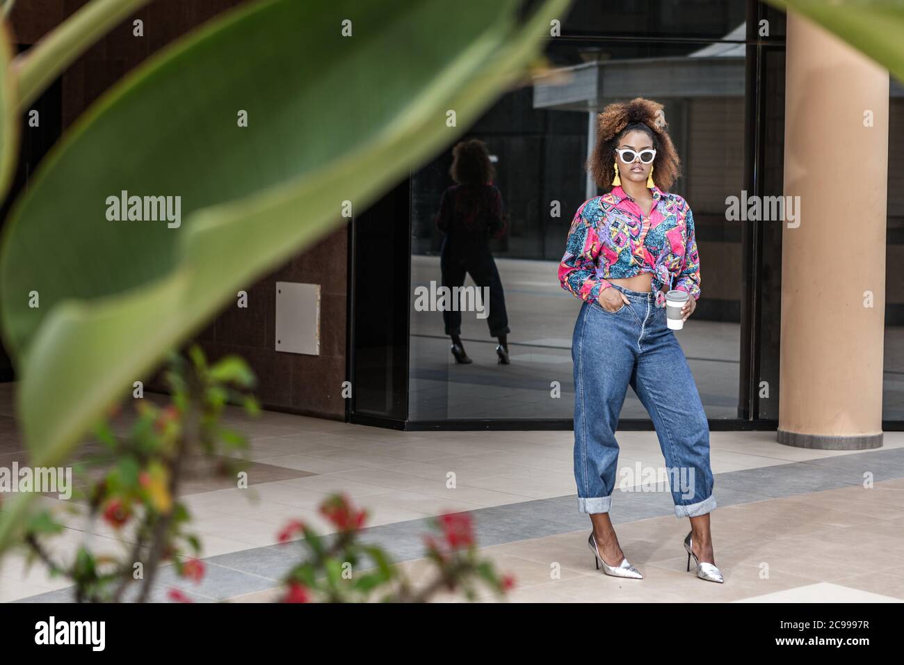 Full body of unemotional black female wearing fashionable clothes and sunglasses standing in city with take away coffee in reusable cup and looking at Stock Photo