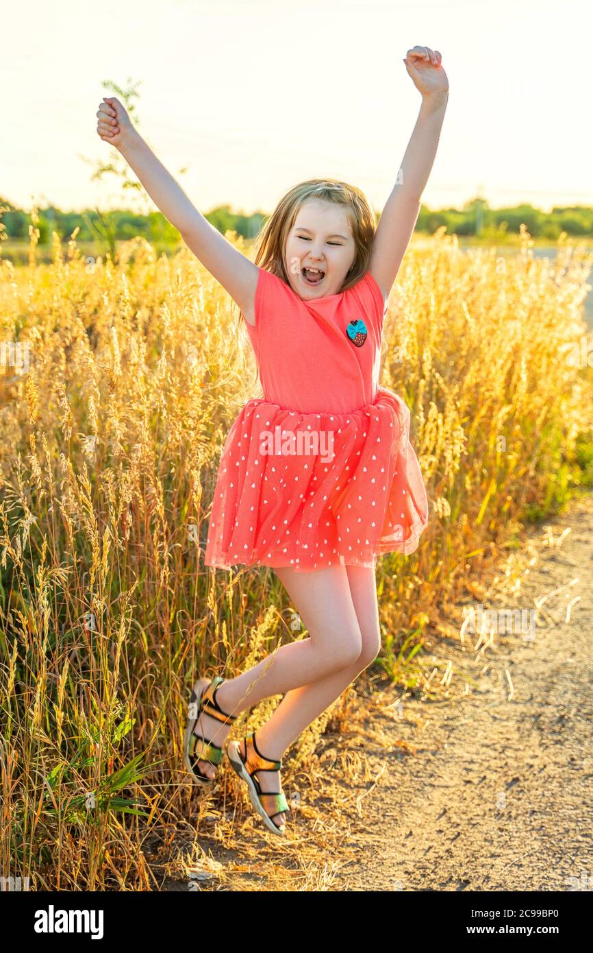 Cute screaming little girl in pink dress jump outdor in field in sunset. Activities with children summer outdoors. Having fun. Vertical. Stock Photo