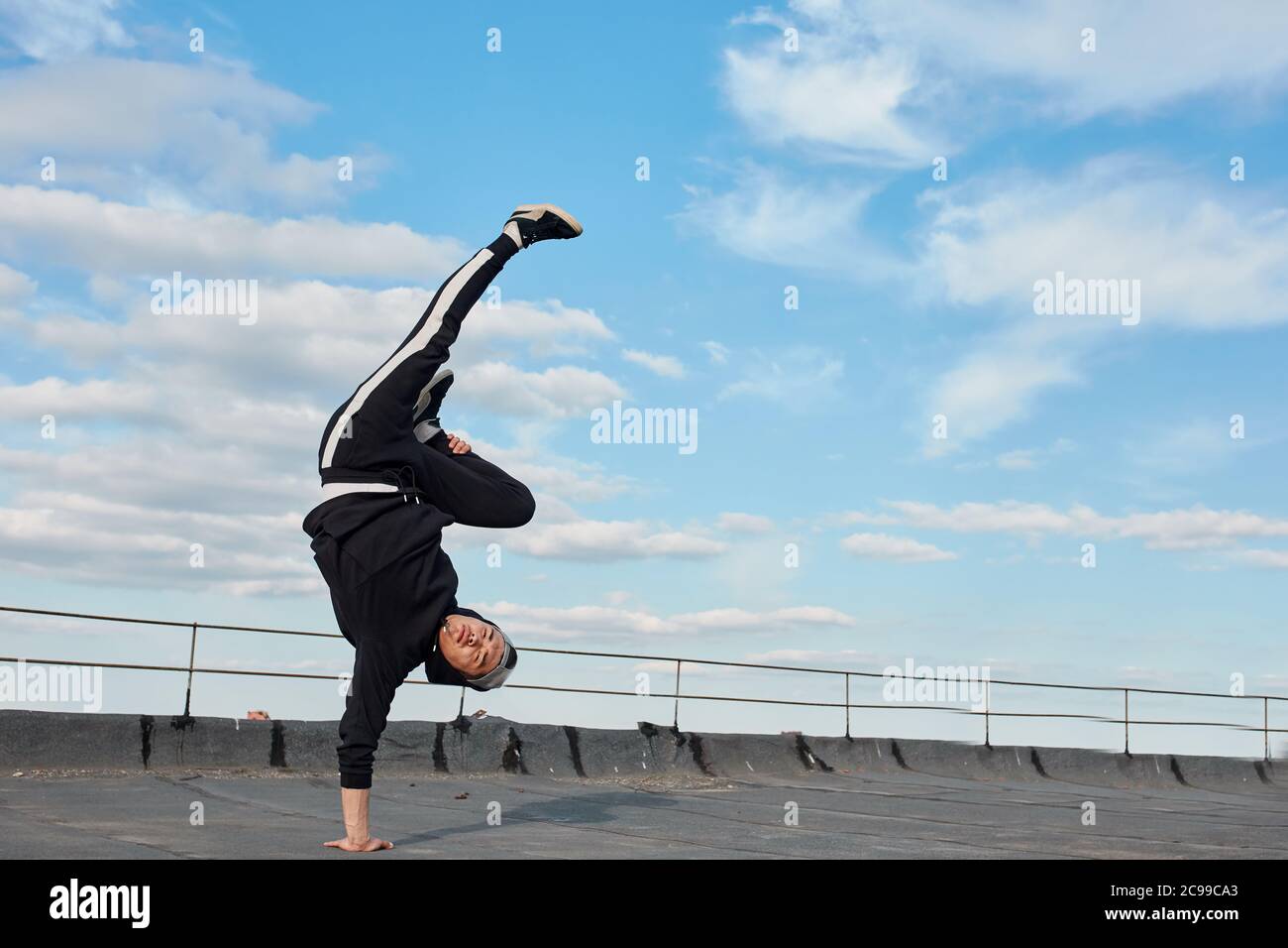 Young Korean urban style male dancer working out, performing breakdance moves, handstand on the top roof outdoor against blue sky. Stock Photo