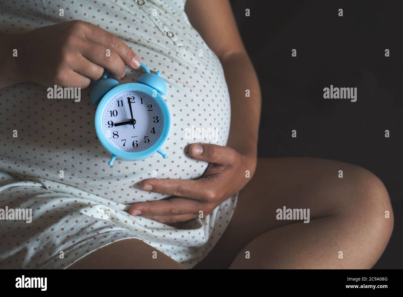 High angle of crop pregnant female holding alarm clock on belly while waiting for baby. Stock Photo