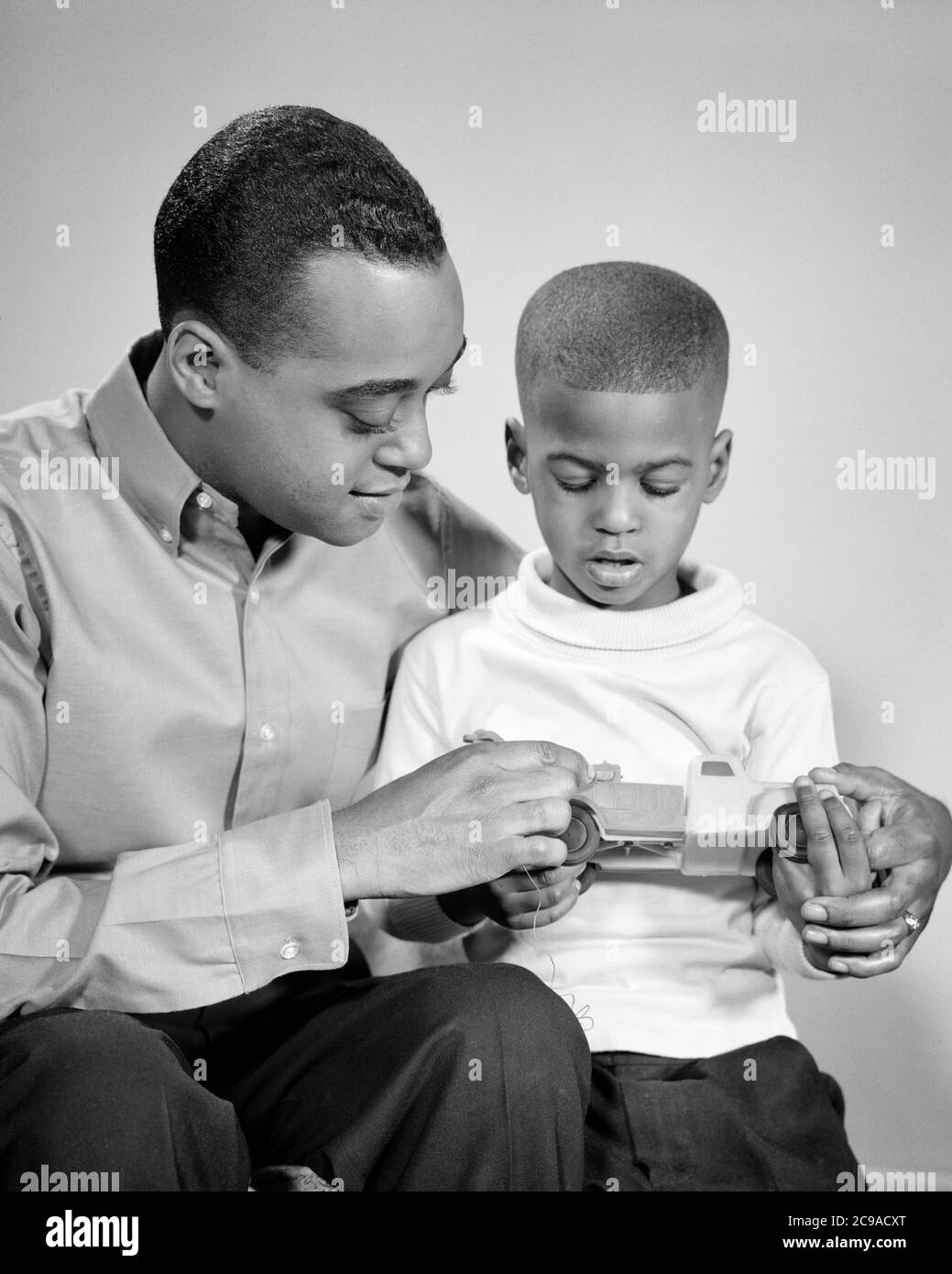 1960s AFRICAN-AMERICAN MAN FATHER WITH SON SITTING SIDE-BY-SIDE HOLDING TOY CAR - n2230 HAR001 HARS COMMUNICATION YOUNG ADULT TEAMWORK SONS FAMILIES JOY LIFESTYLE SATISFACTION STUDIO SHOT HOME LIFE COPY SPACE FRIENDSHIP HALF-LENGTH PERSONS INSPIRATION MALES FATHERS B&W HAPPINESS DISCOVERY EXPLAINING HIS AFRICAN-AMERICANS AFRICAN-AMERICAN DADS BLACK ETHNICITY SUPPORT GROWTH JUVENILES SIDE-BY-SIDE TOGETHERNESS YOUNG ADULT MAN BLACK AND WHITE HAR001 OLD FASHIONED AFRICAN AMERICANS Stock Photo