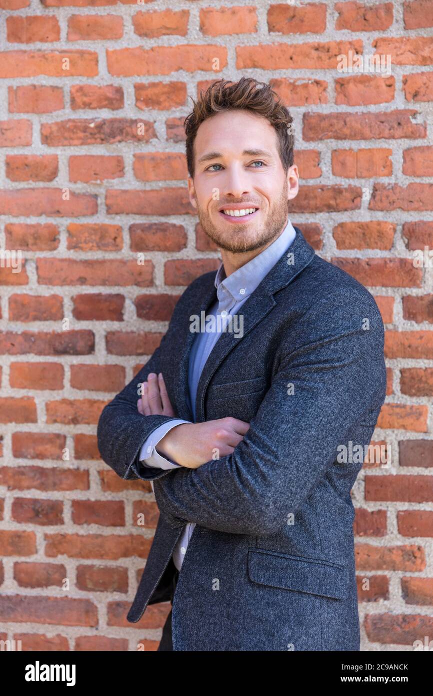 Smart casual businessman on urban city brick wall background lifestyle portrait. Young professional man smiling confident in blazer. Career and Stock Photo