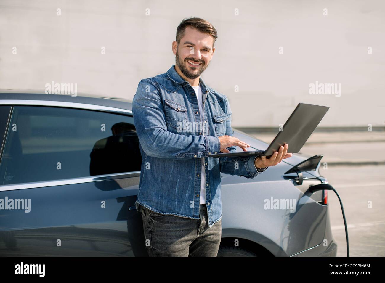 Young handsome smiling man working on laptop, waiting while his luxury electric car is charching, paying for charging, standing near city station with Stock Photo