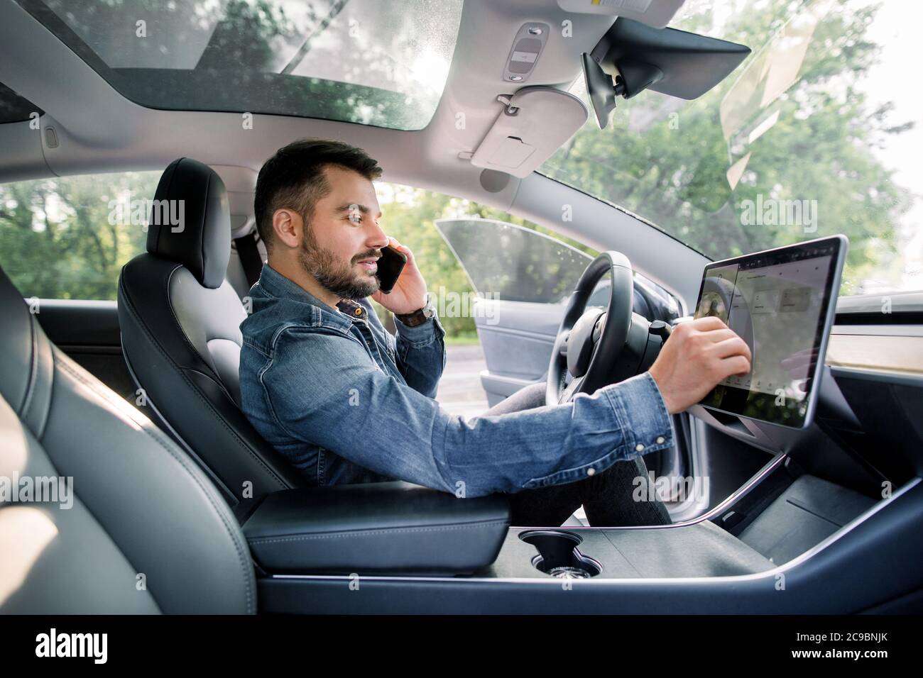 Testing a new electric futuristic car with self driving system. Side view of satisfied Caucasian man in casual jeans shirt sitting in modern car Stock Photo