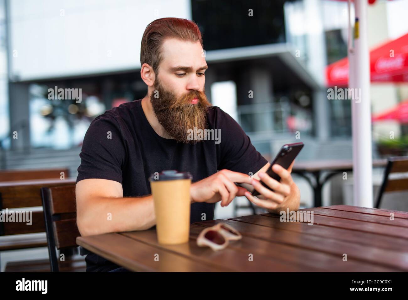 Portrait of young man with the beard texting message into his workgroup using the smartphone while waiting for a food order in street restaurant Stock Photo