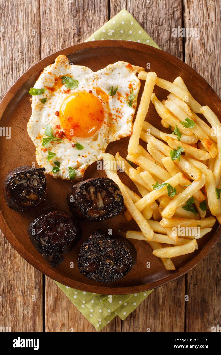 Huevos rotos con morcilla fried egg with fries and blood sausage close-up in a plate on the table. Vertical top view from above Stock Photo