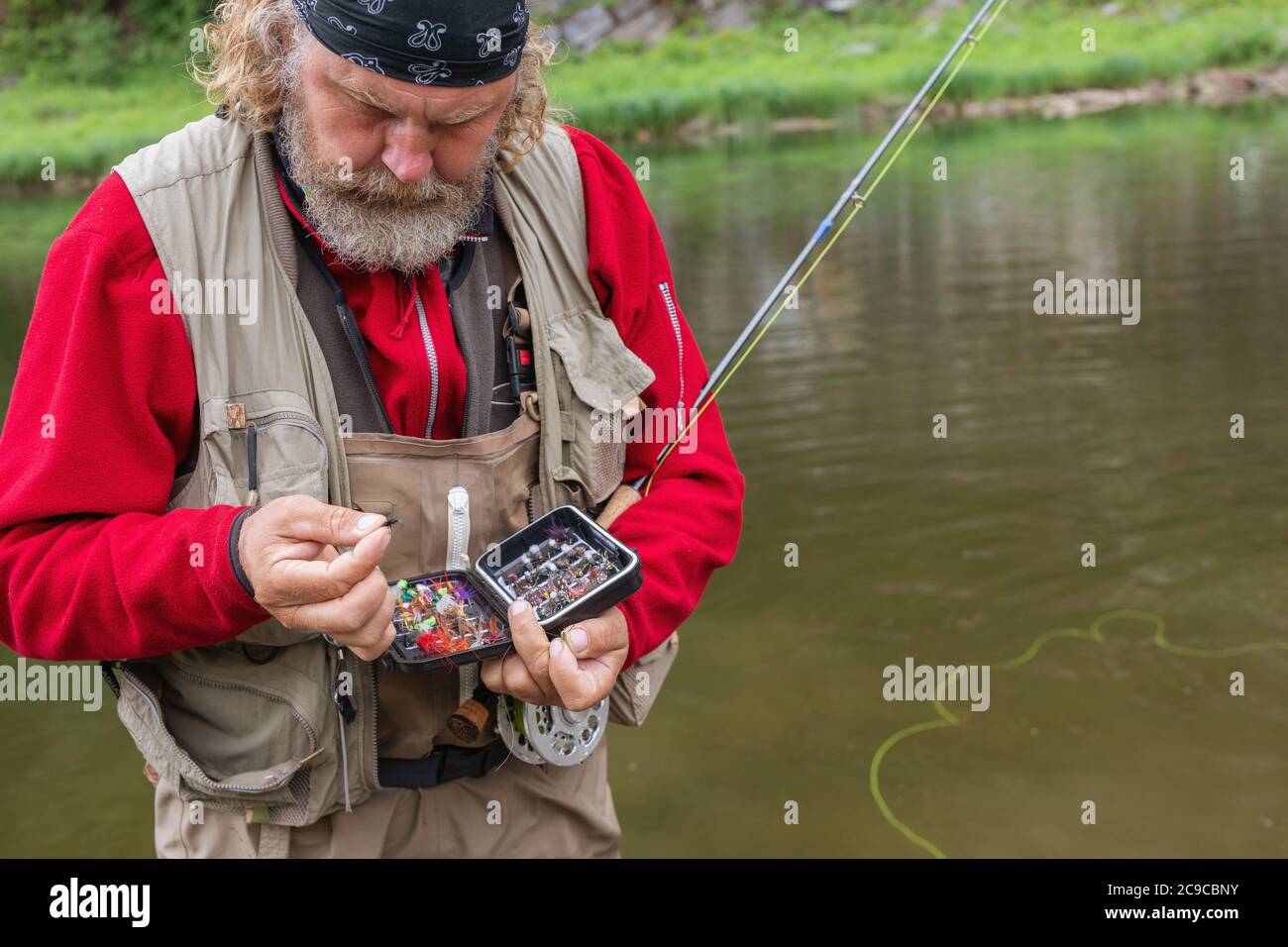 Serious elderly bearded fisherman on river bank holding rod and tackle box  selects fly fishing lure would be better. Copy space. Fly-fishing, active l  Stock Photo - Alamy
