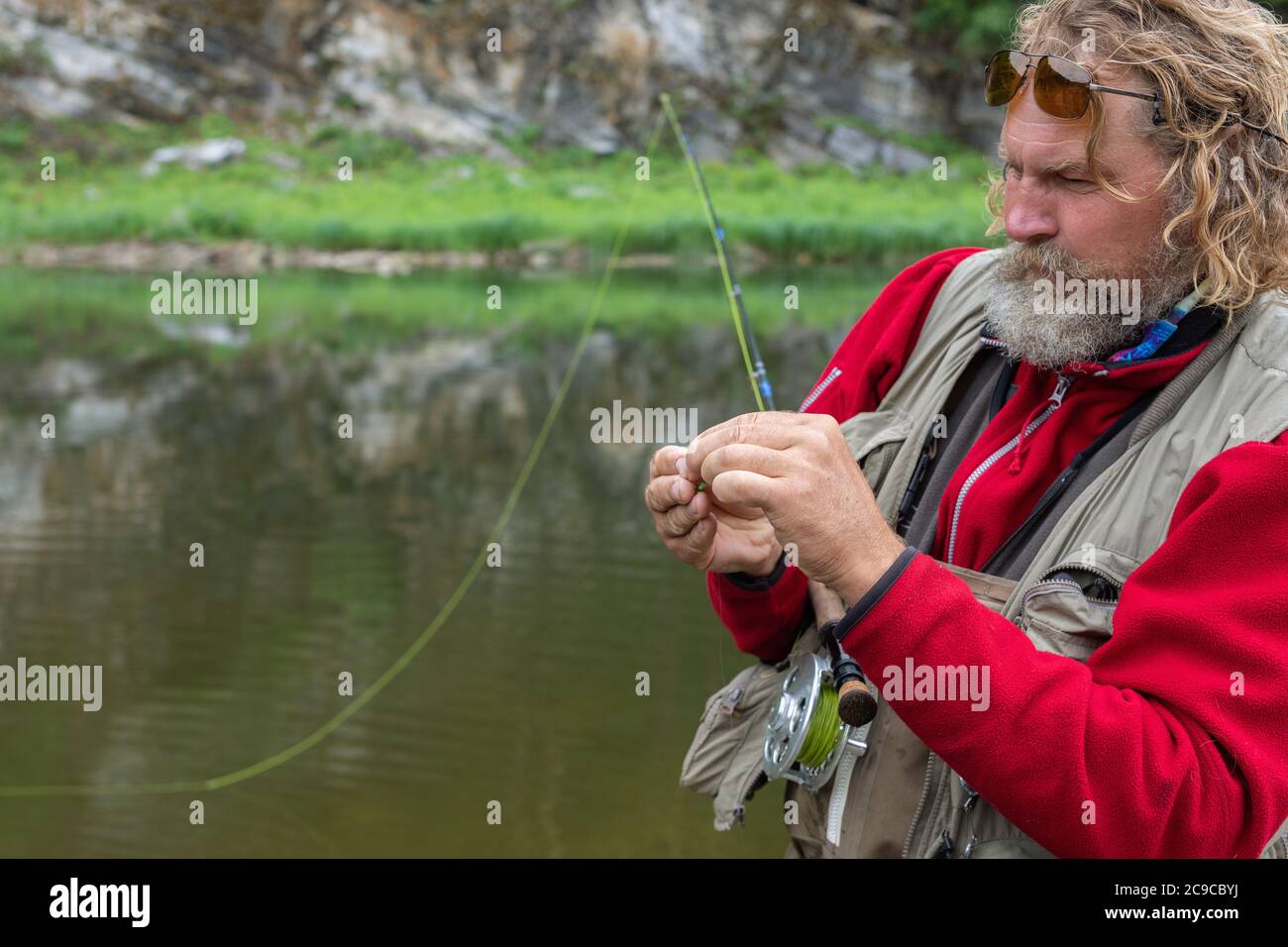 Mature fly fishing fisherman in polarizing glasses tying fly of fishing hook on fishing rod Stock Photo