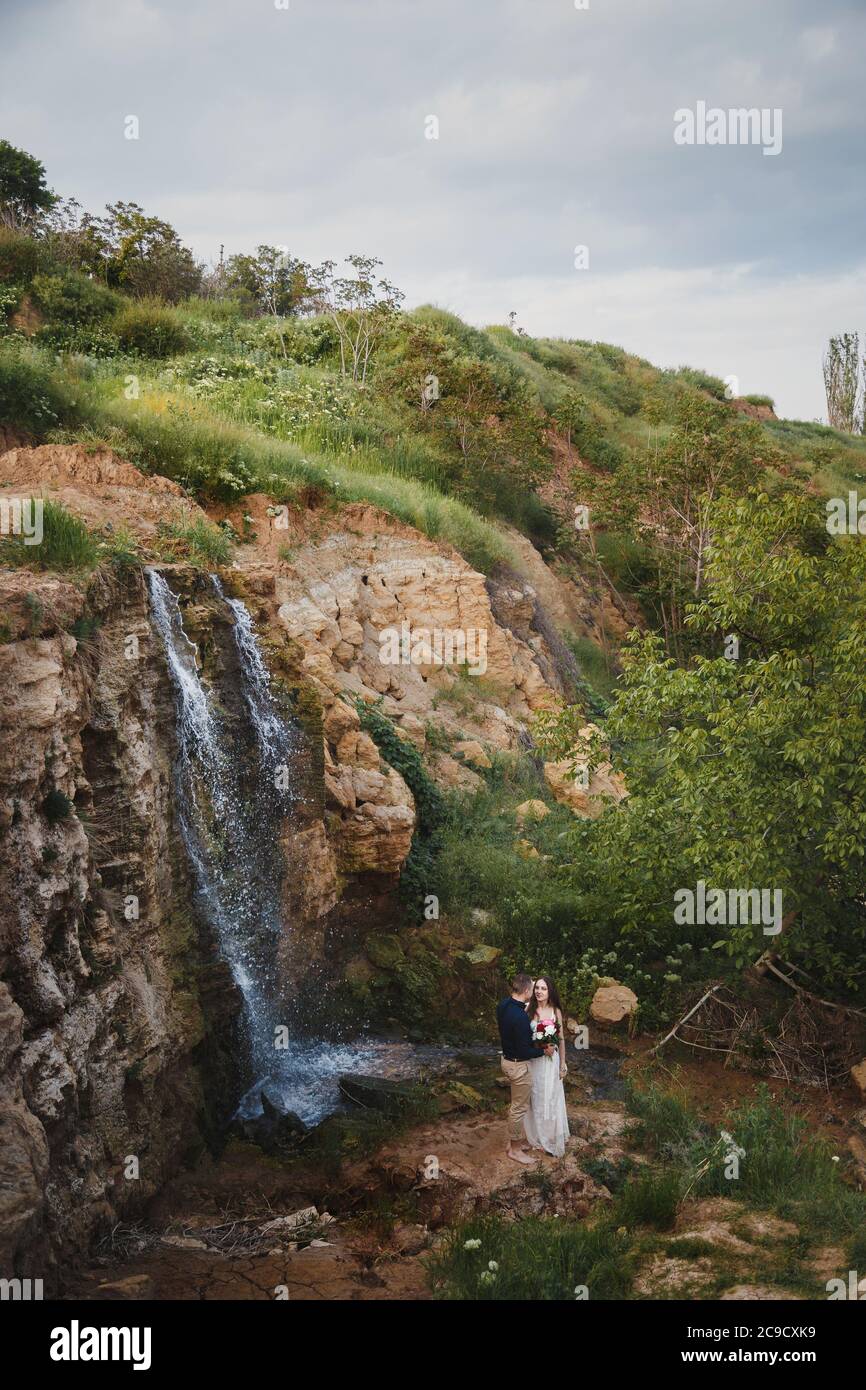 Outdoor wedding ceremony, stylish happy smiling groom and bride are hugging and kissing in front of small waterfall Stock Photo