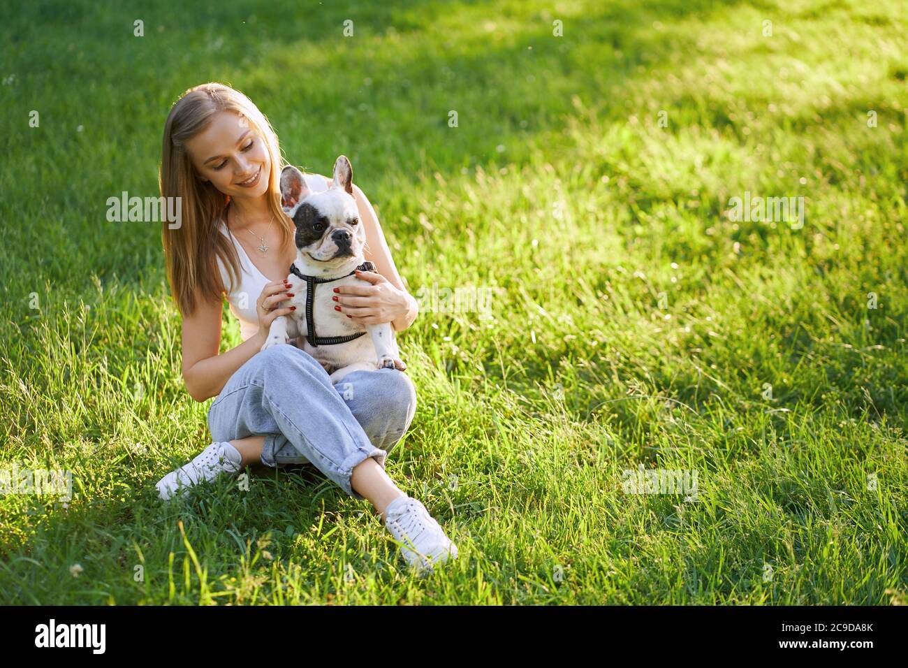Top view of young happy woman sitting on grass with friendly french bulldog. Gorgeous caucasian smiling girl enjoying summer sunset, holding dog on knees and showing love and care in city park. Stock Photo