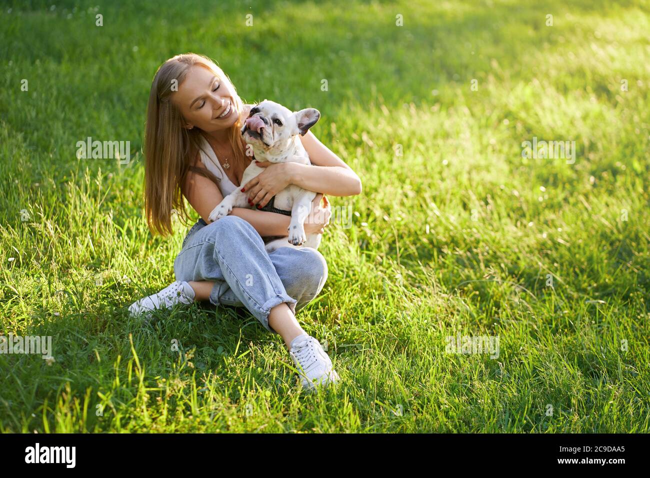 Top view of young happy woman sitting on grass with friendly french bulldog. Gorgeous caucasian smiling girl enjoying summer sunset, holding dog on knees and showing love and care in city park. Stock Photo