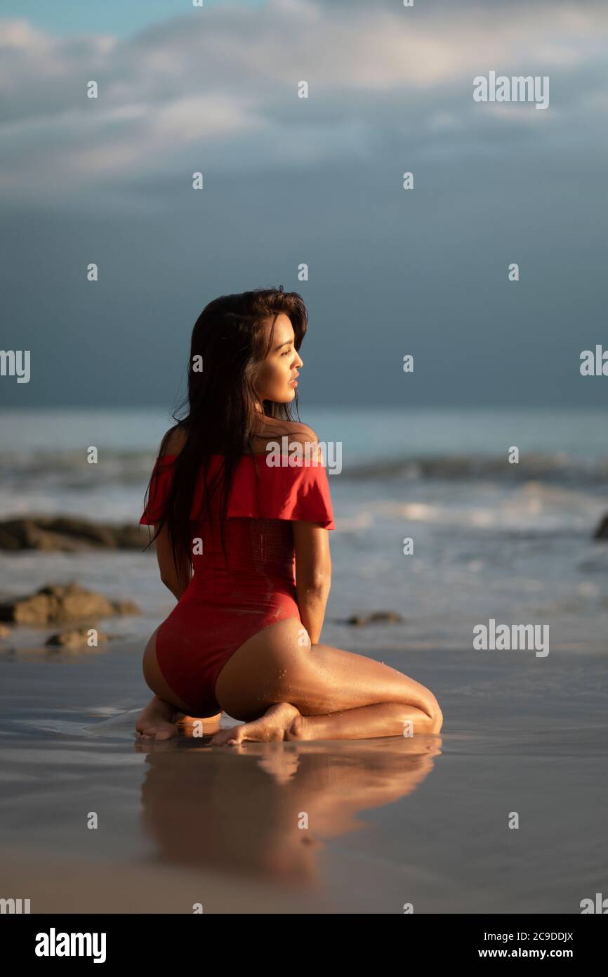 Backside view of beautiful Mexican woman in red swimsuit sitting at a beach looking into the sunset Stock Photo