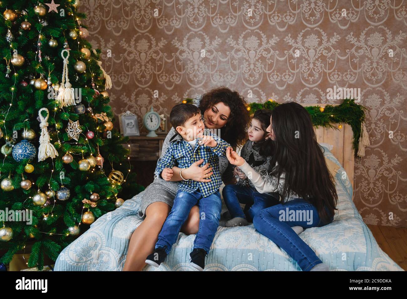 Christmas family portrait of happy smiling women sitting on bed hugging and playing with their small children near to christmas tree at home. Winter Stock Photo