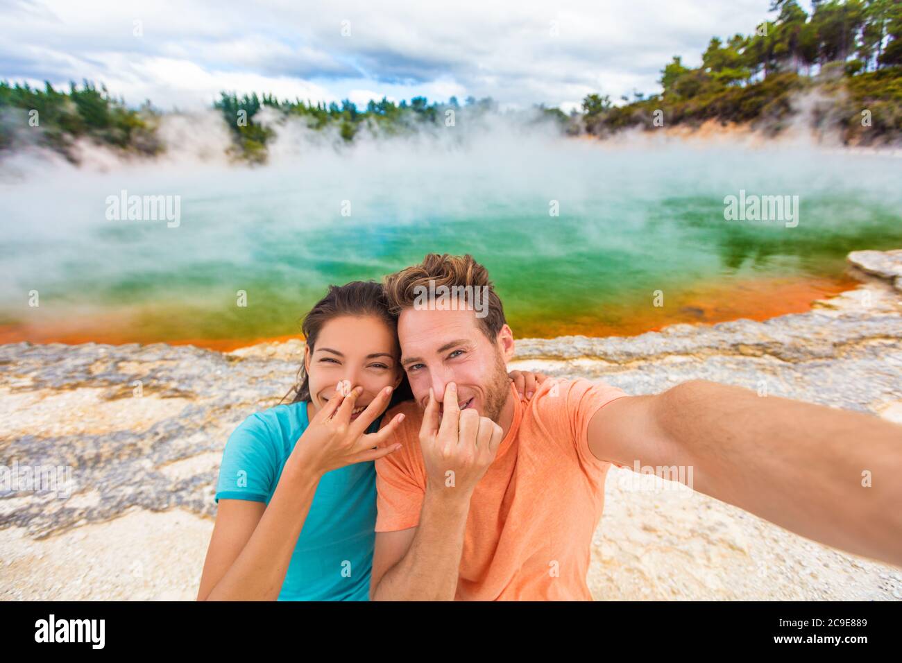 Funny selfie couple tourists at New Zealand pools travel. Young people doing goofy face at smell bad sulfur smell at colorful geothermal hot springs Stock Photo