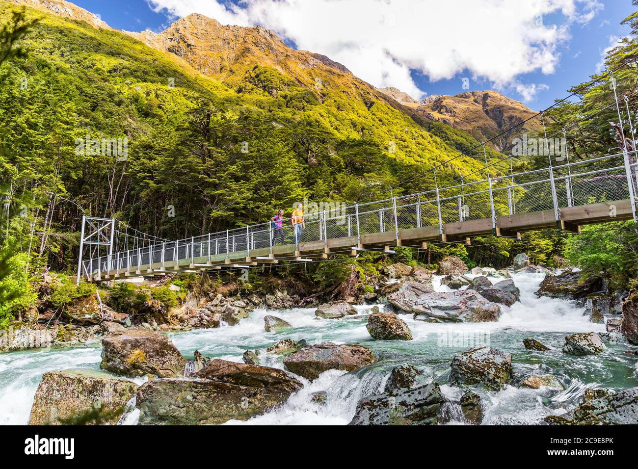 New Zealand tramping people crossing river bridge. Hikers couple backpackers walking hiking together with backpacks on Routeburn Track trail path. Stock Photo