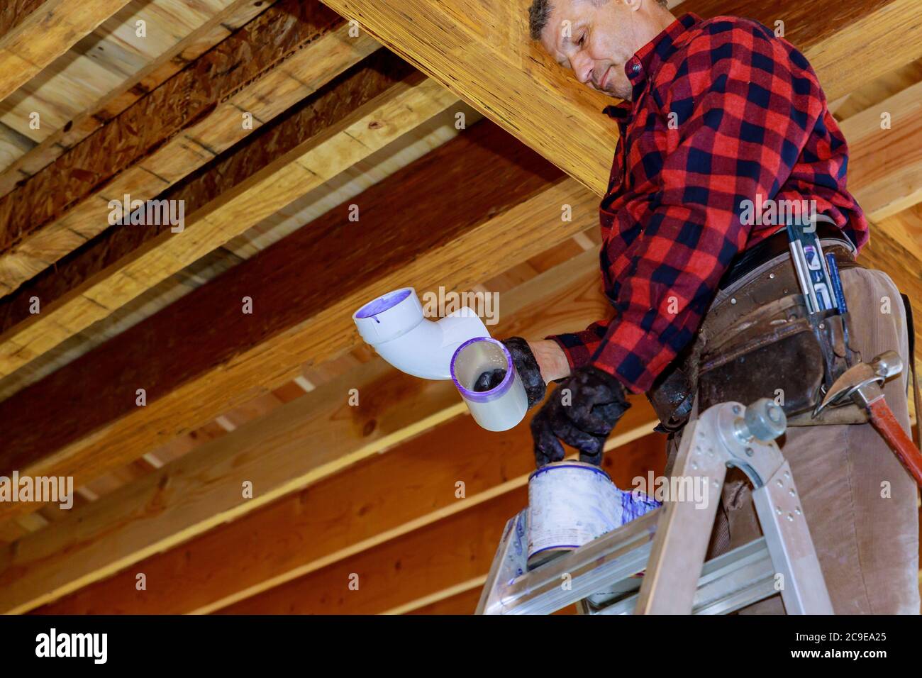 Plumber gluing PVC plastic sewer toilet white pipe on wooden frame ceiling beam of house soft focus Stock Photo