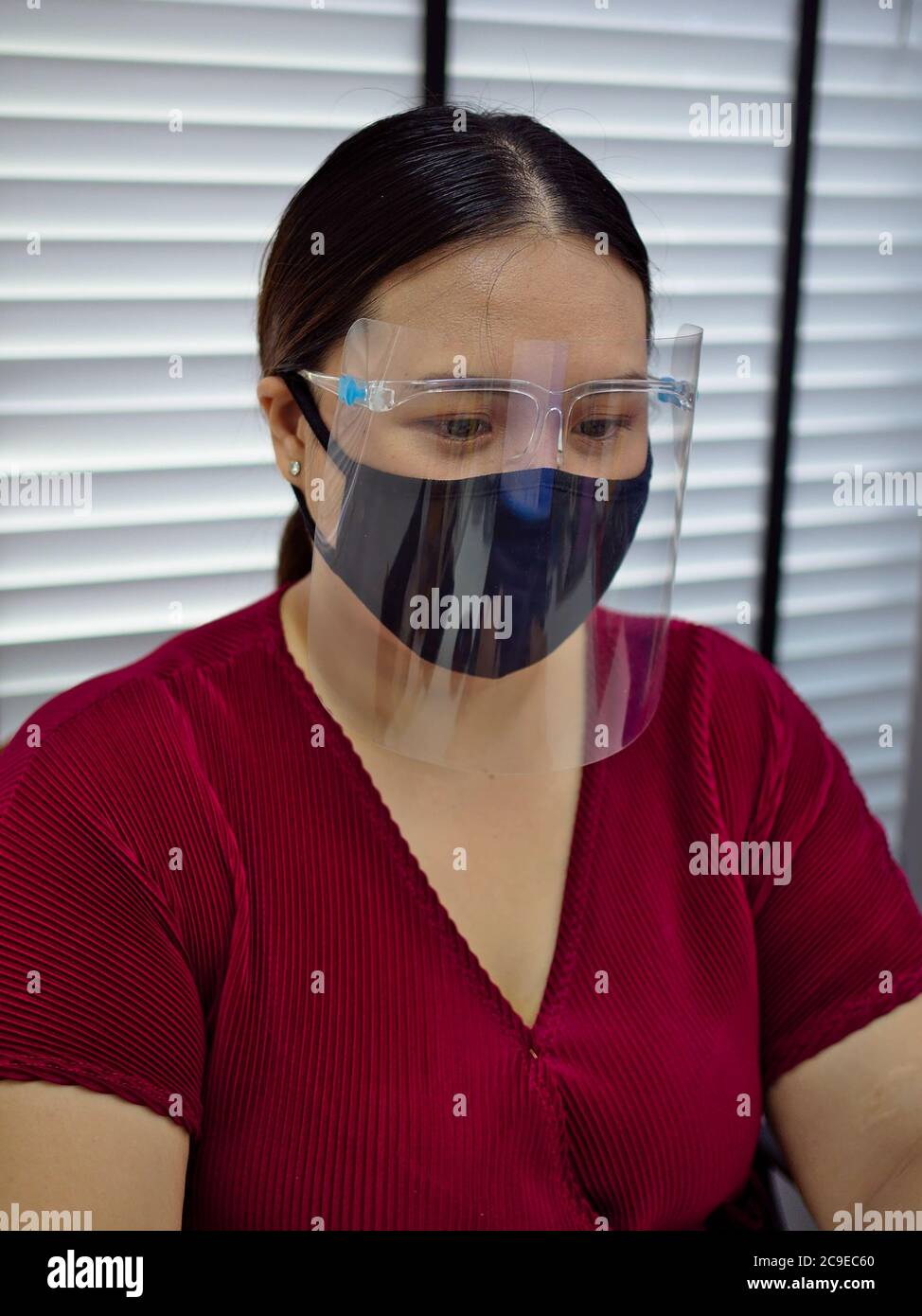 A young and beautiful Asian woman wearing a surgical mask and face shield to protect against Covid-19 while working in an office. Stock Photo