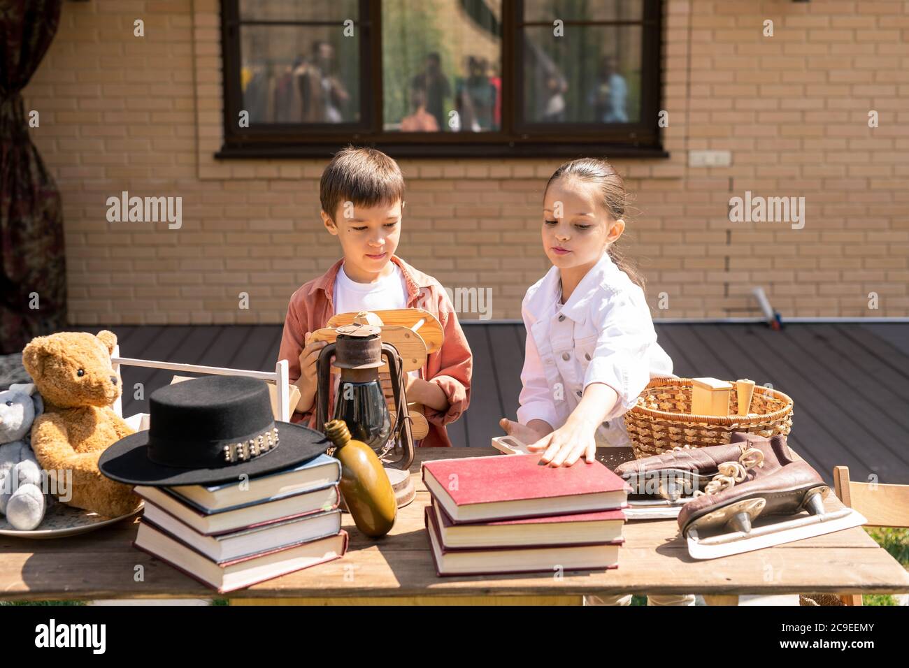 Cute children standing at table with various goods and buying toy and book at garage sale Stock Photo