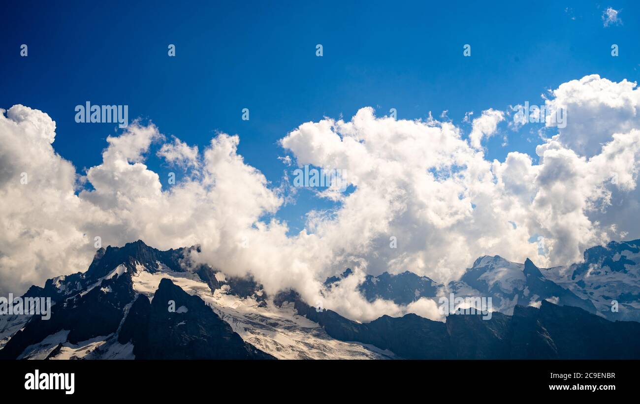 Snowy mountain against cloudy sky. From below white clouds floating on blue sky over mountain ridge covered with snow on sunny day in nature Stock Photo