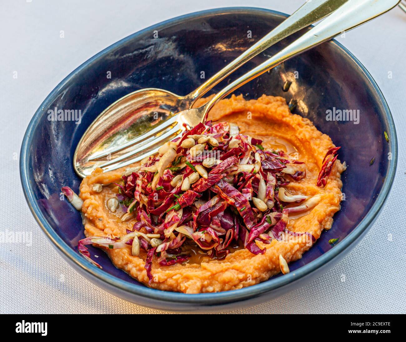 A dark blue bowl holds "Crema di Fagioli", a creamy purée of giant beans, topped with a radicchio and sunflower-seed salad, with silver cutlery laid ready beside it. In 2020, during the coronavirus pandemic, the La Cucina restaurant at Hotel Mühle in Binzen in the Markgräflerland region, Baden-Württemberg, Germany, switched from its previous Markgräfler cuisine to Apulian dishes for the first time. Stock Photo