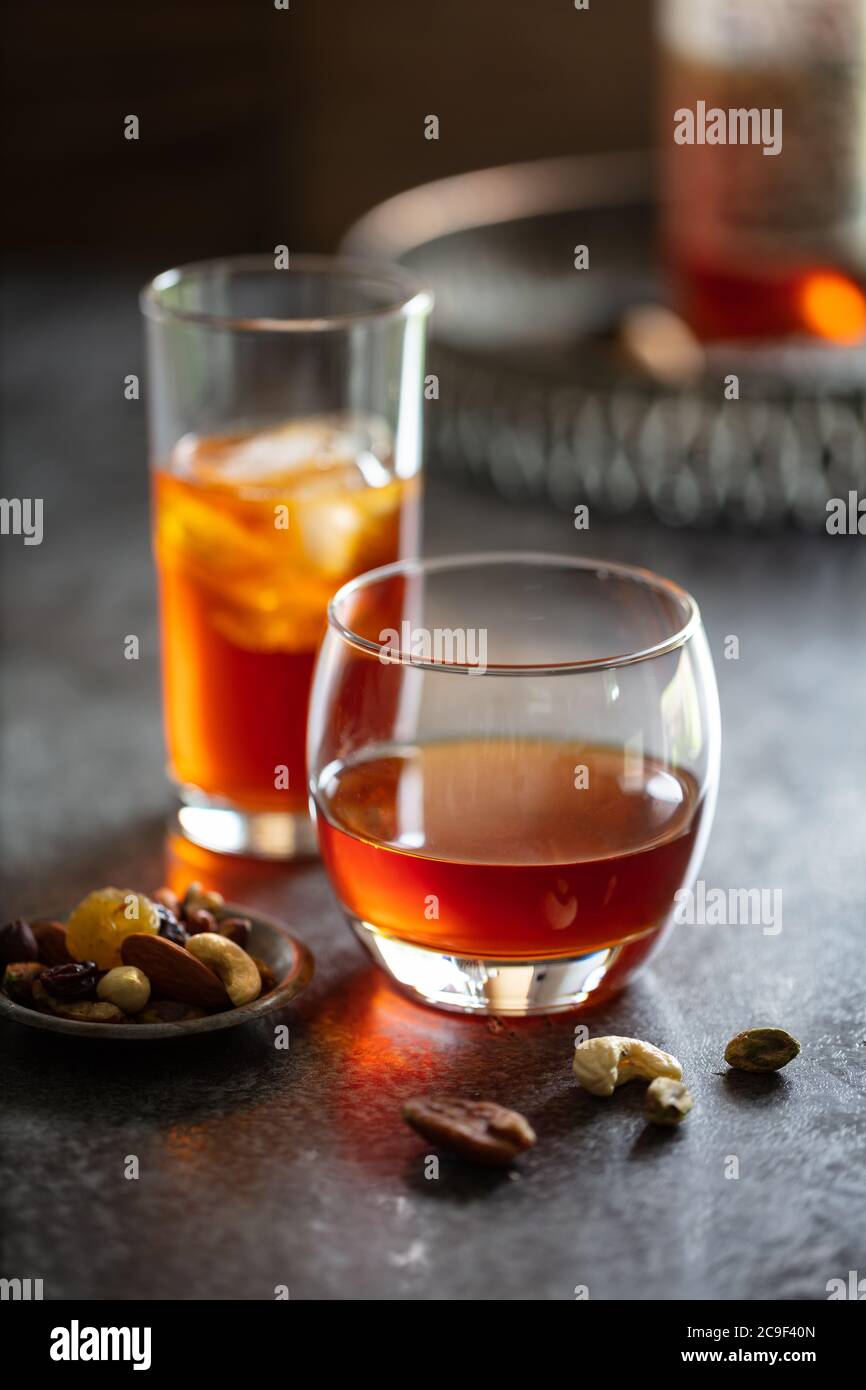 Alcoholic drinks (whiskey vermouth) with ice on dark worktop.  Dish of nibbles. Portrait Stock Photo