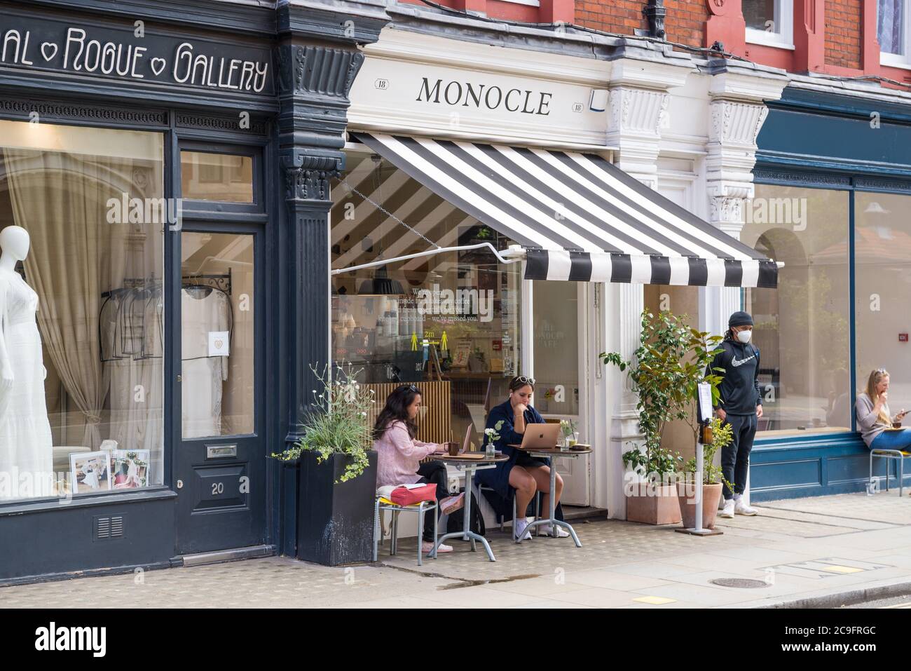 Two young women seated at pavement tables outside the Monocle Cafe taking refreshment and using laptop computers. Chiltern Street, Marylebone, London Stock Photo