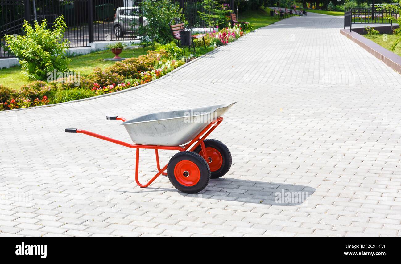 Side view on a metallic wheelbarrow with red wheels standing on paved path in blooming public park Stock Photo