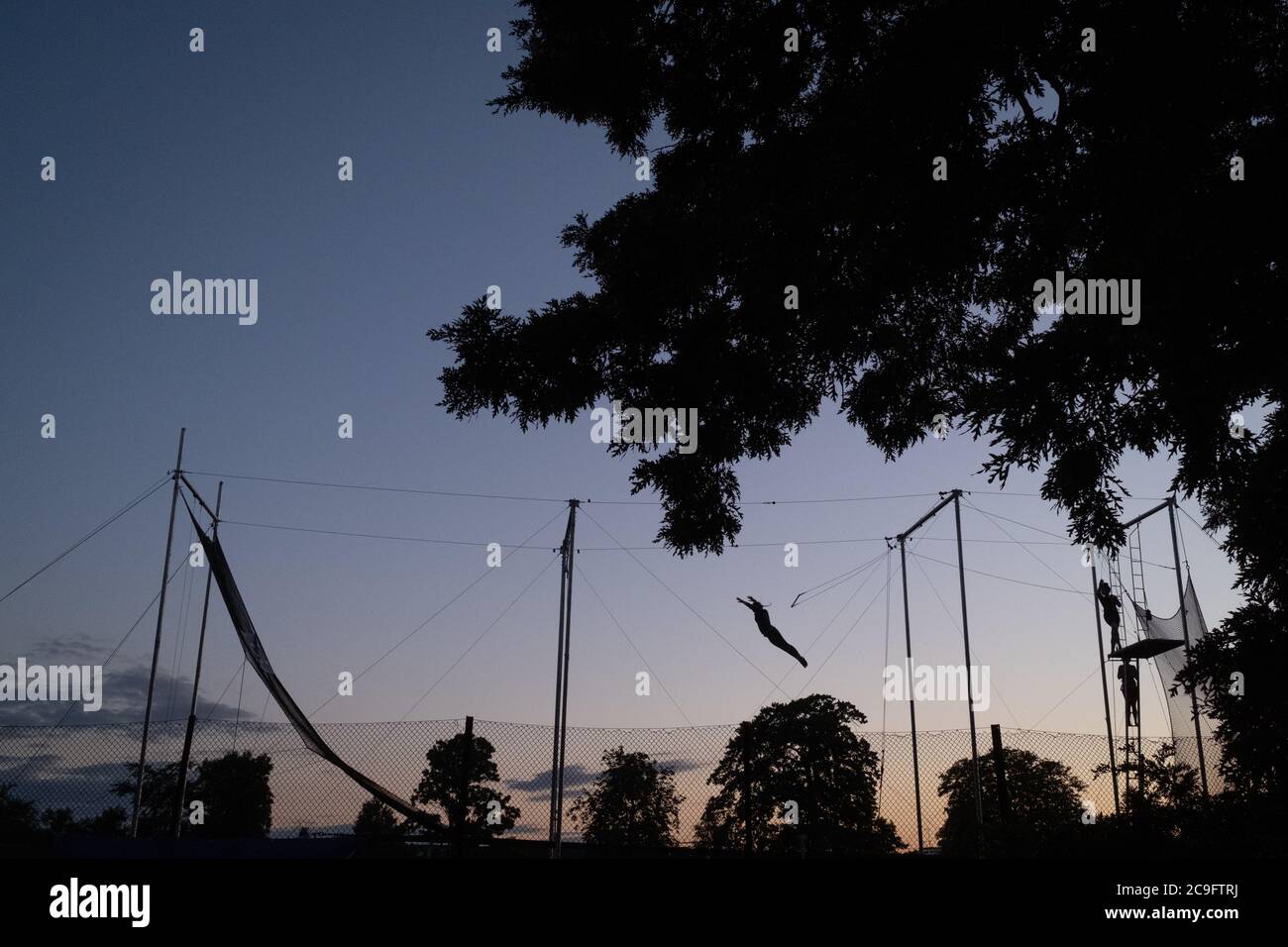 Silhouetted against evening skies, a figure performs trapeze acrobatics in Ruskin Park in Lambeth, on 29th July 2020, in London, England. TLCC Trapeze School comes to Ruskin Park each summer although during the Coronavirus pandemic, they have had to delay their presence in this south London Park. Stock Photo