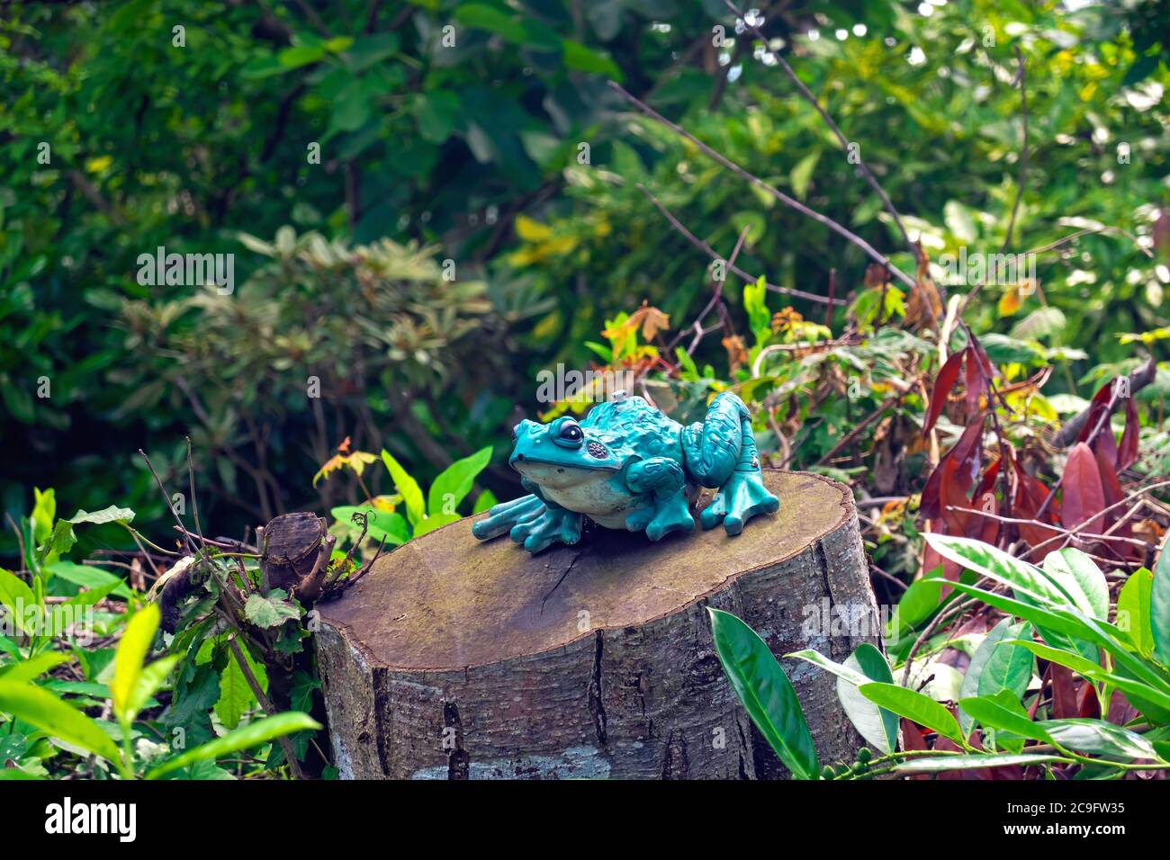 An ornamental turquoise garden frog sitting on a tree stump with shrubs in the background. Stock Photo