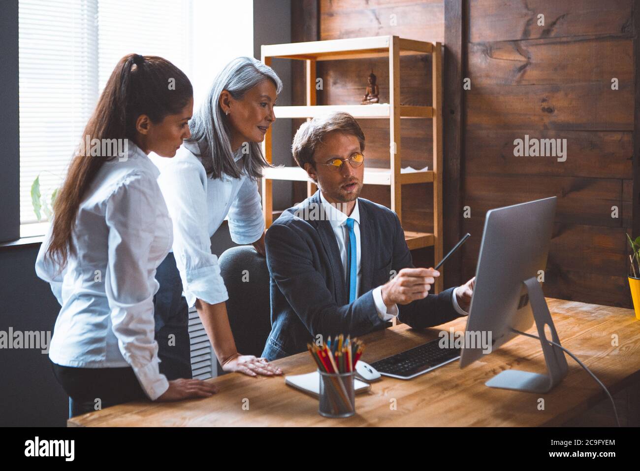 Team work of diverse office team. Young brunette and gray haired woman listen their male colleague pointing to computer monitor. Coworking concept Stock Photo