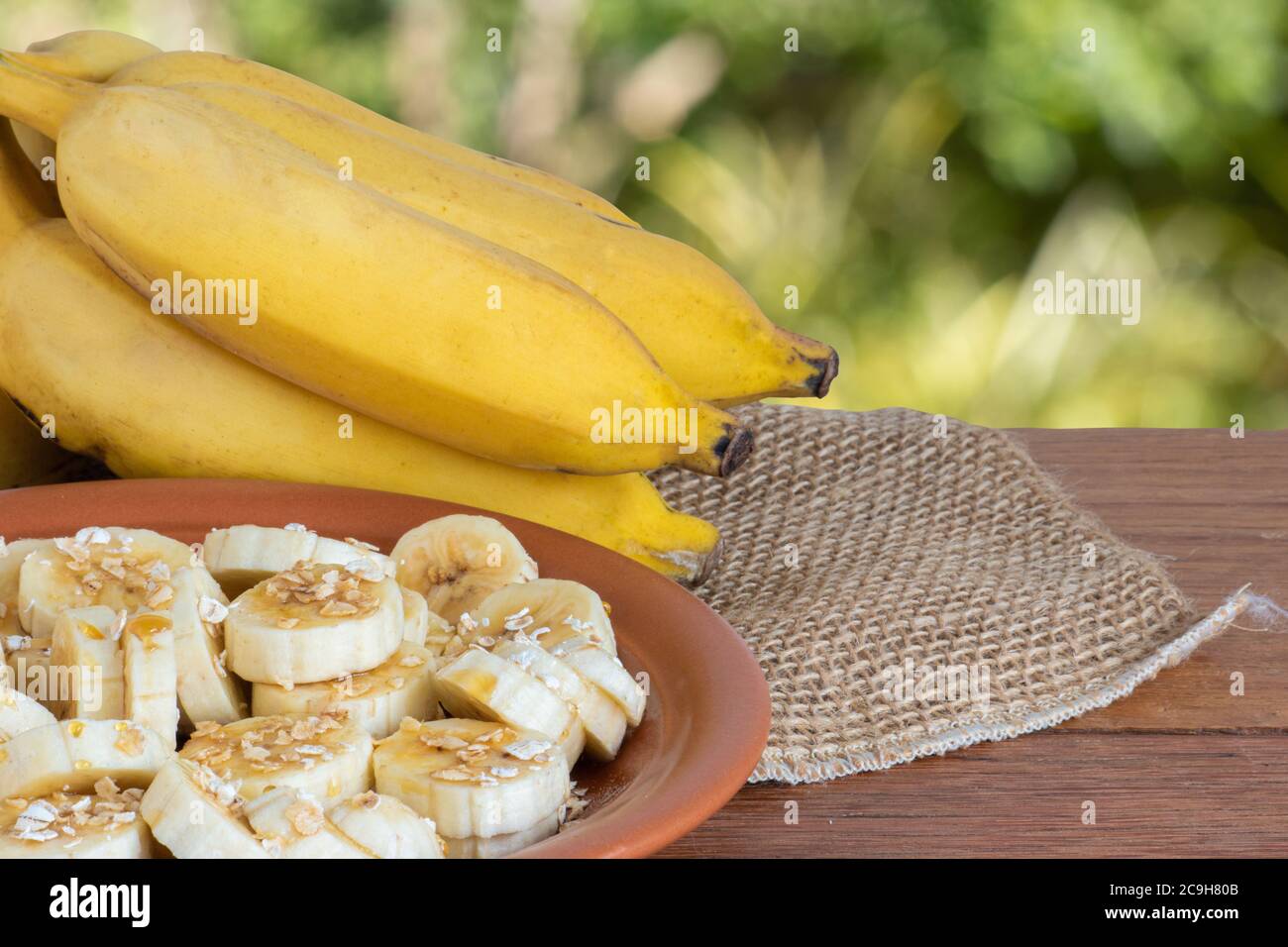 Organic bunch of ripe yellow bananas. Pieces cut on a plate with honey and oats. Isolated with blurred green background.Copy space Stock Photo