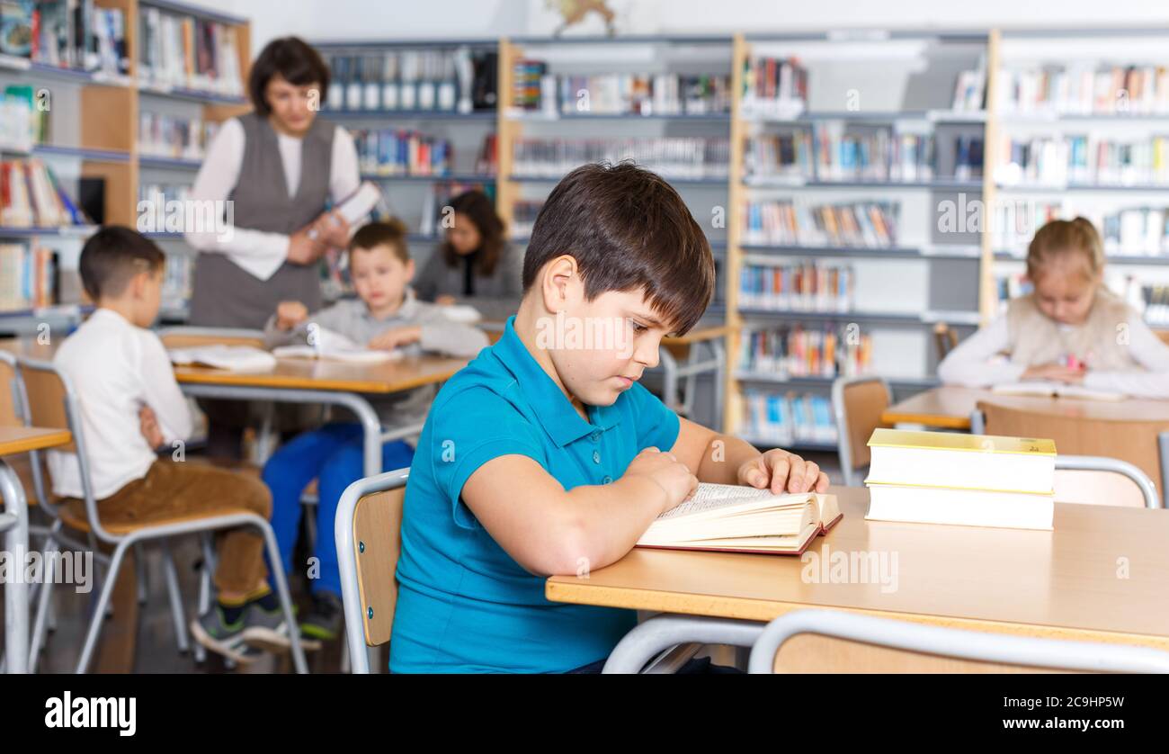 Focused boy reading in school library on background with other students and teacher Stock Photo