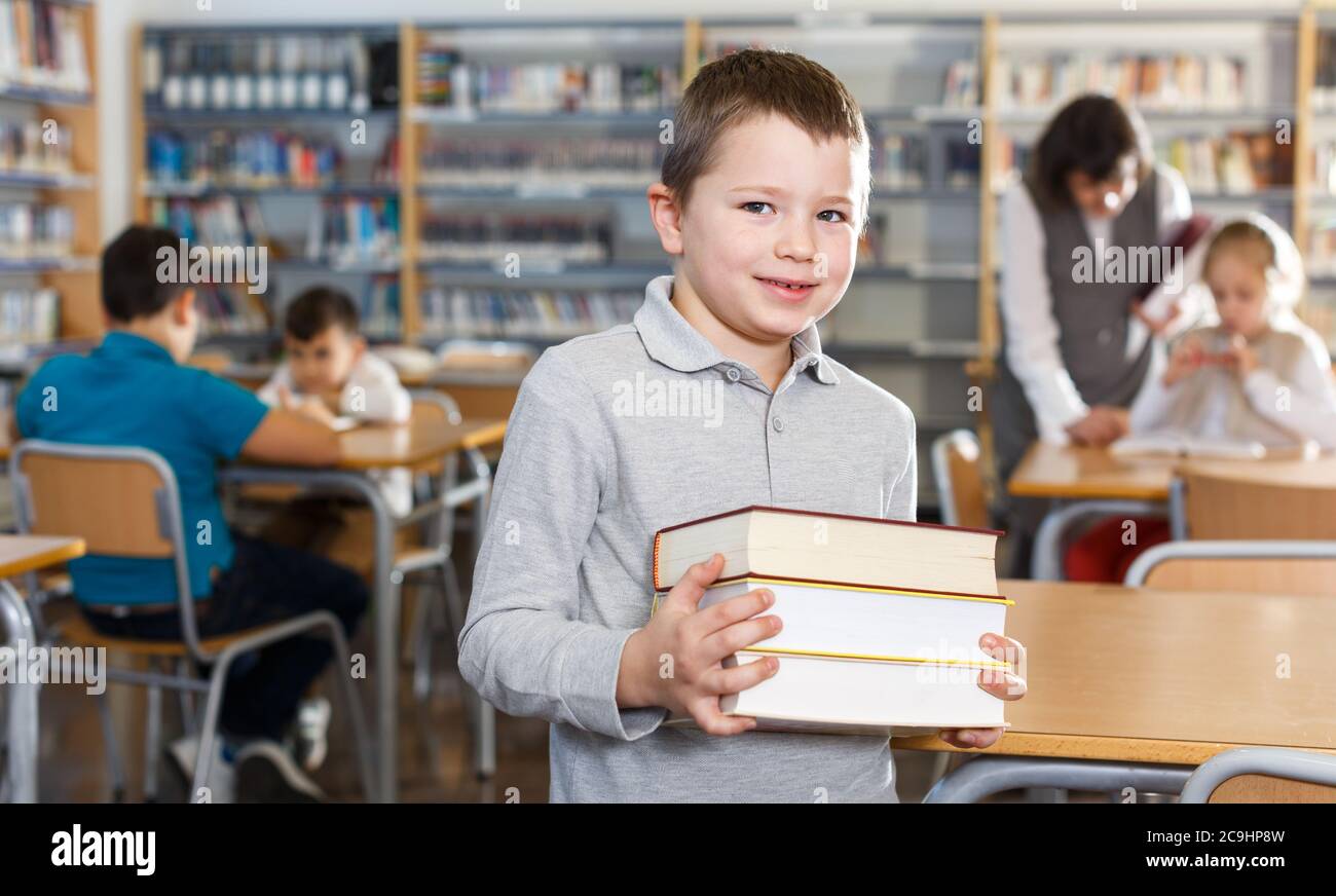 Portrait of smiling preteen boy standing with pile of books in school ...