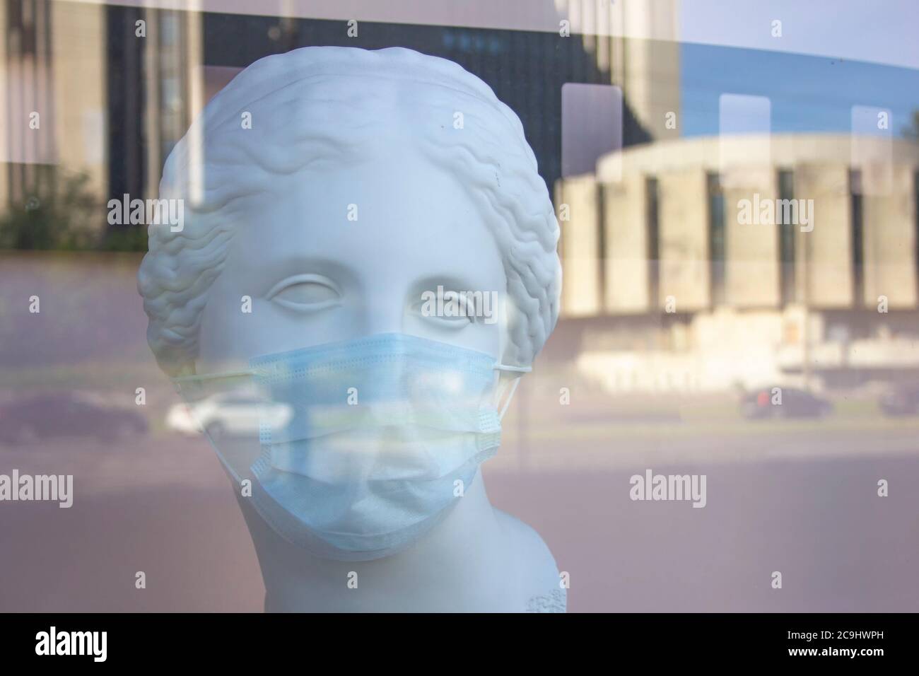 a man, woman plaster bust in a protective medical mask behind a plate-glass window with reflection of city streets Stock Photo
