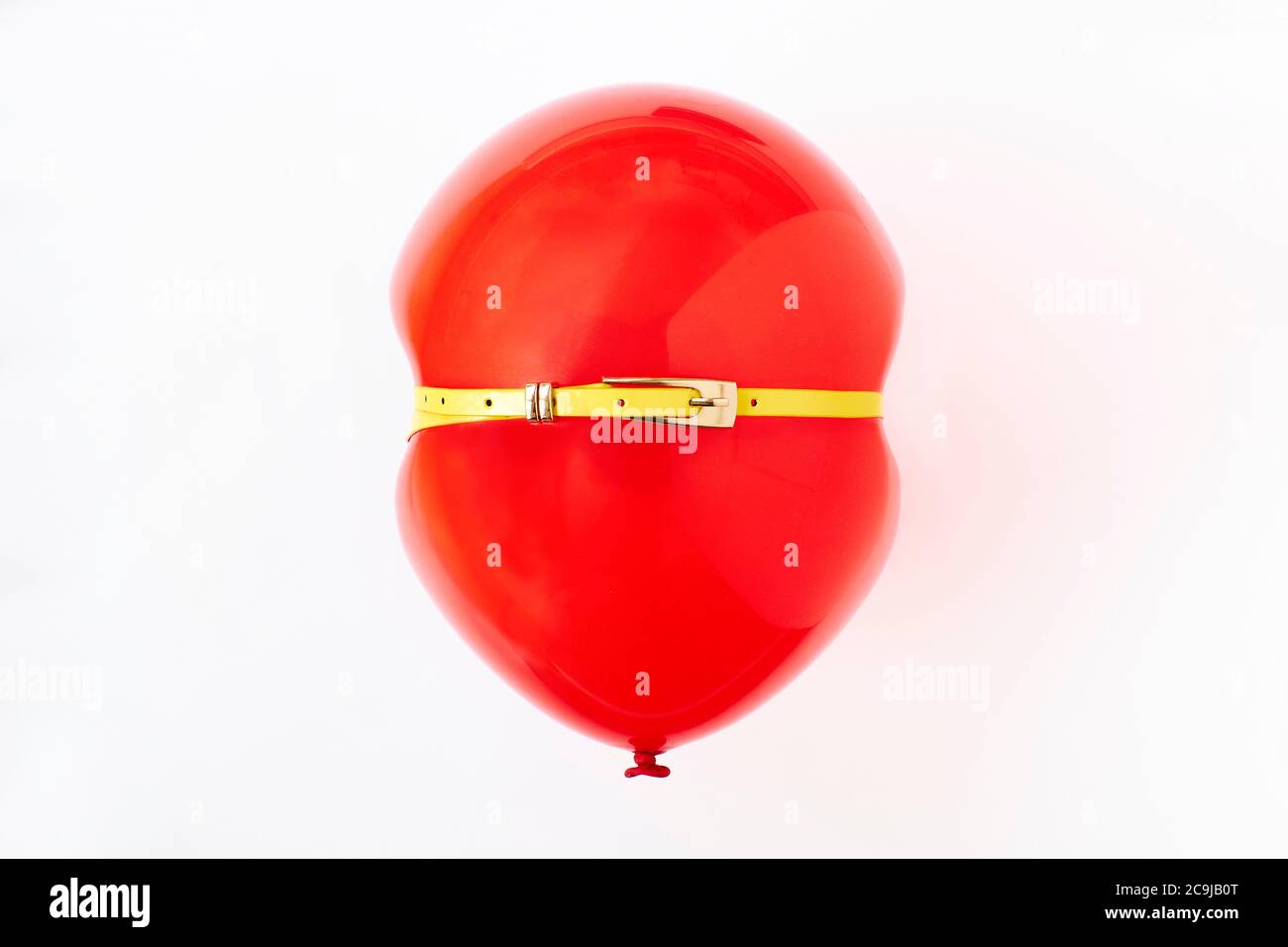 Red balloon squeezed by yellow belt, conceptual image of bloated stomach. Stock Photo