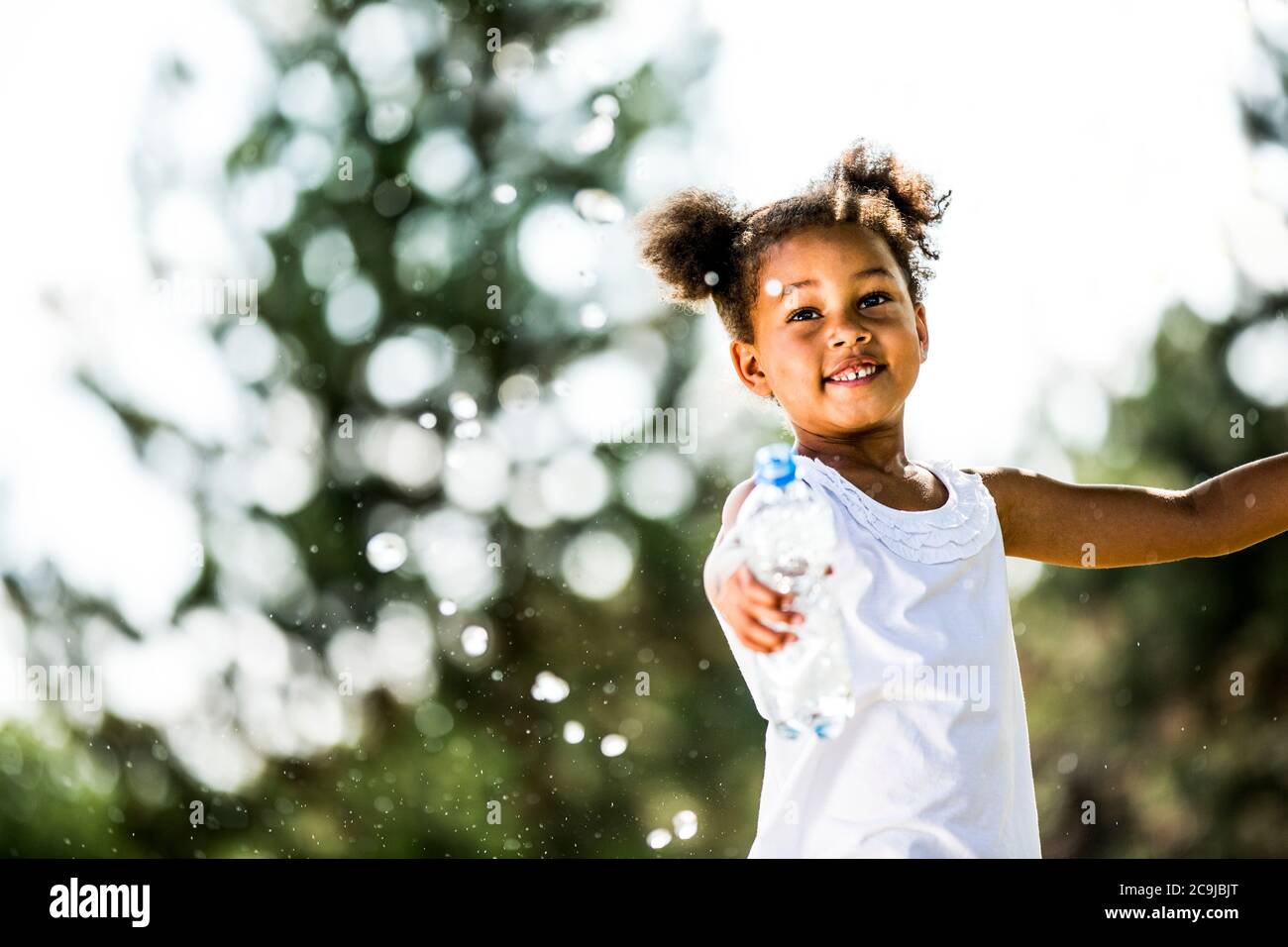 Girl with bunches splashing water from plastic bottle, smiling. Stock Photo