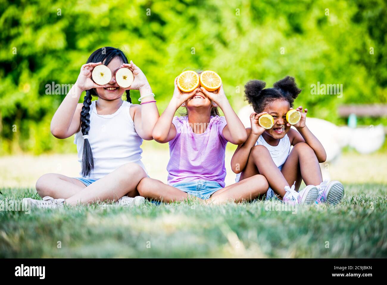 Girls sitting side by side in park and covering their eyes with fruit slices, smiling. Stock Photo