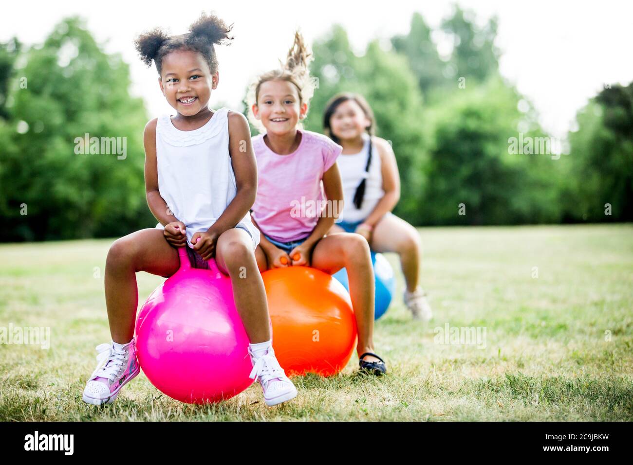 Girls bouncing on inflatable hopper in park, smiling. Stock Photo