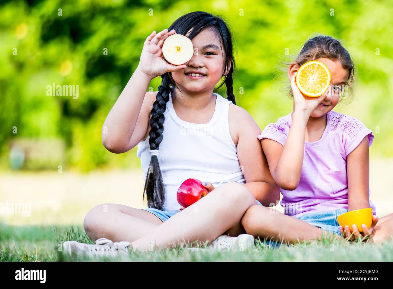 Girls sitting side by side in park and holding various fruits, smiling, portrait. Stock Photo