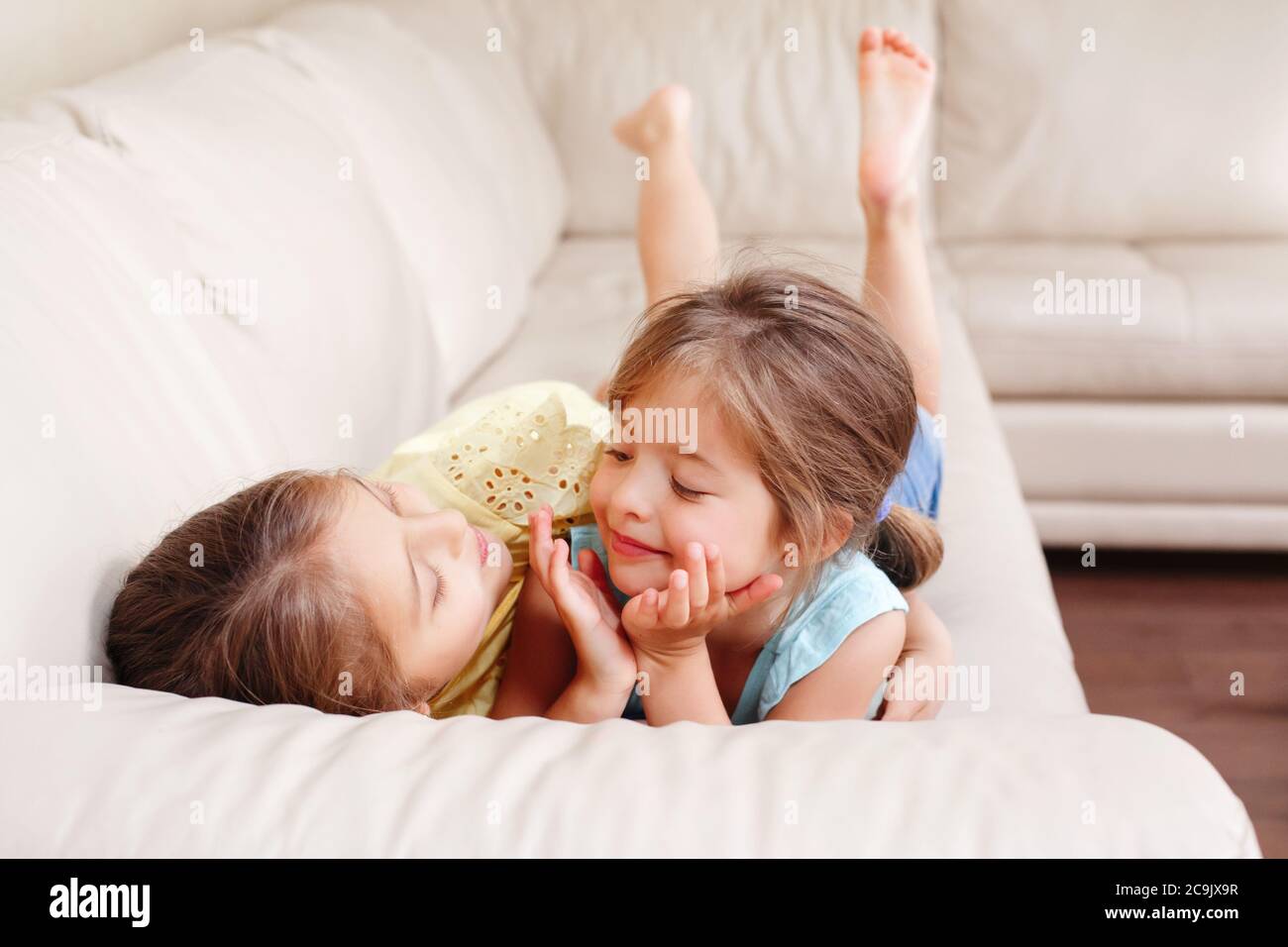 Two cute little Caucasian girls siblings playing at home. Adorable smiling children kids lying on couch together. Authentic candid lifestyle domestic Stock Photo