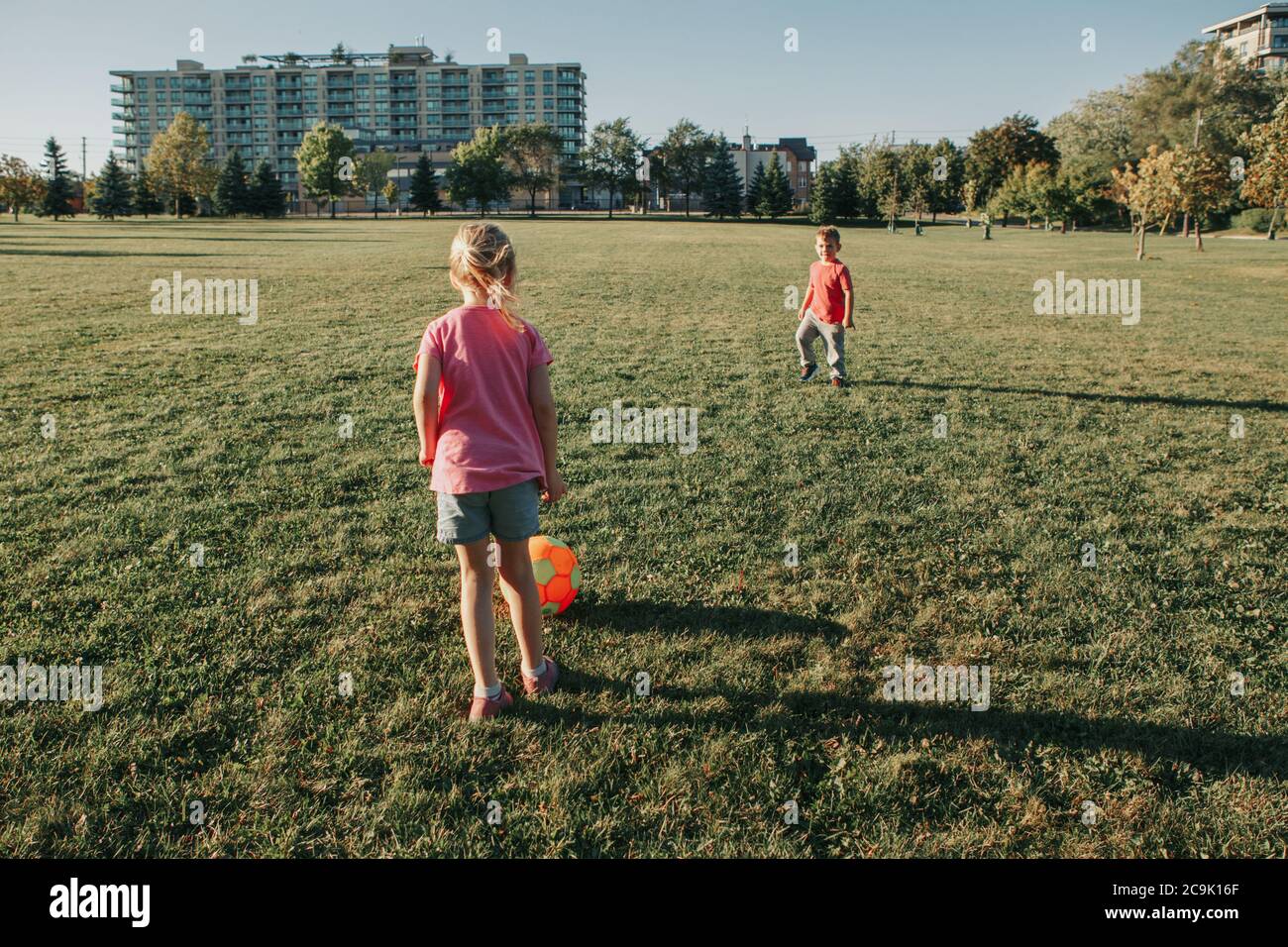 Little preschool girl and boy friends playing soccer football on playground grass field outside. Happy authentic candid childhood lifestyle. Stock Photo