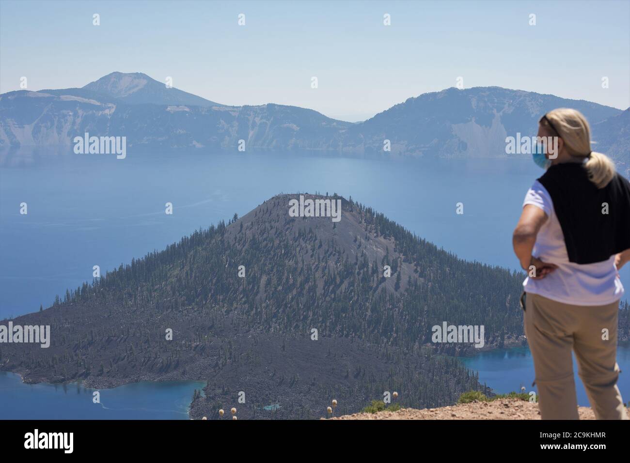 A woman wearing a face mask looks out over Crater Lake and Wizard Island, in Crater Lake National Park in Oregon, USA. Stock Photo