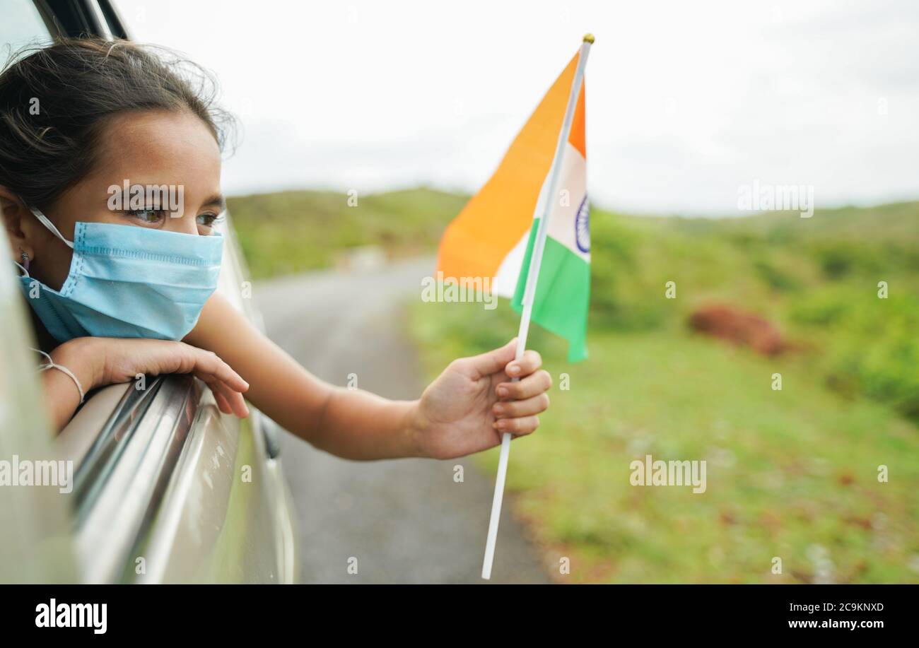 Young girl kid with medical mask holding Indian flag in moving car window - Concept of celebrating Independence or republic day during coronavirus or Stock Photo