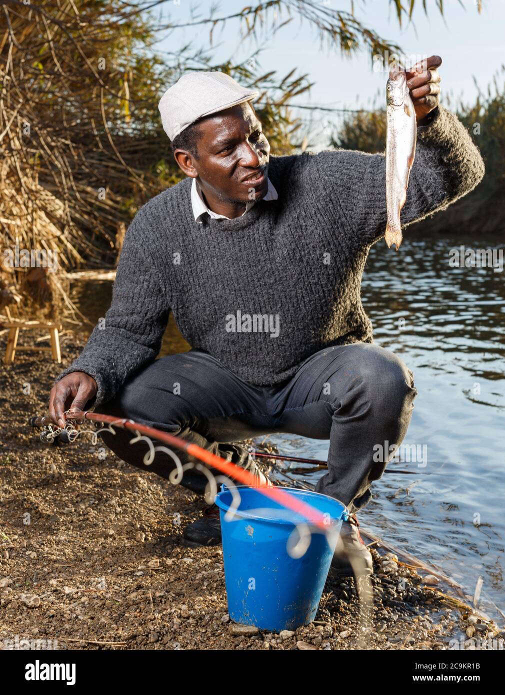 Adult African fisherman putting caught fish in bucket near river Stock